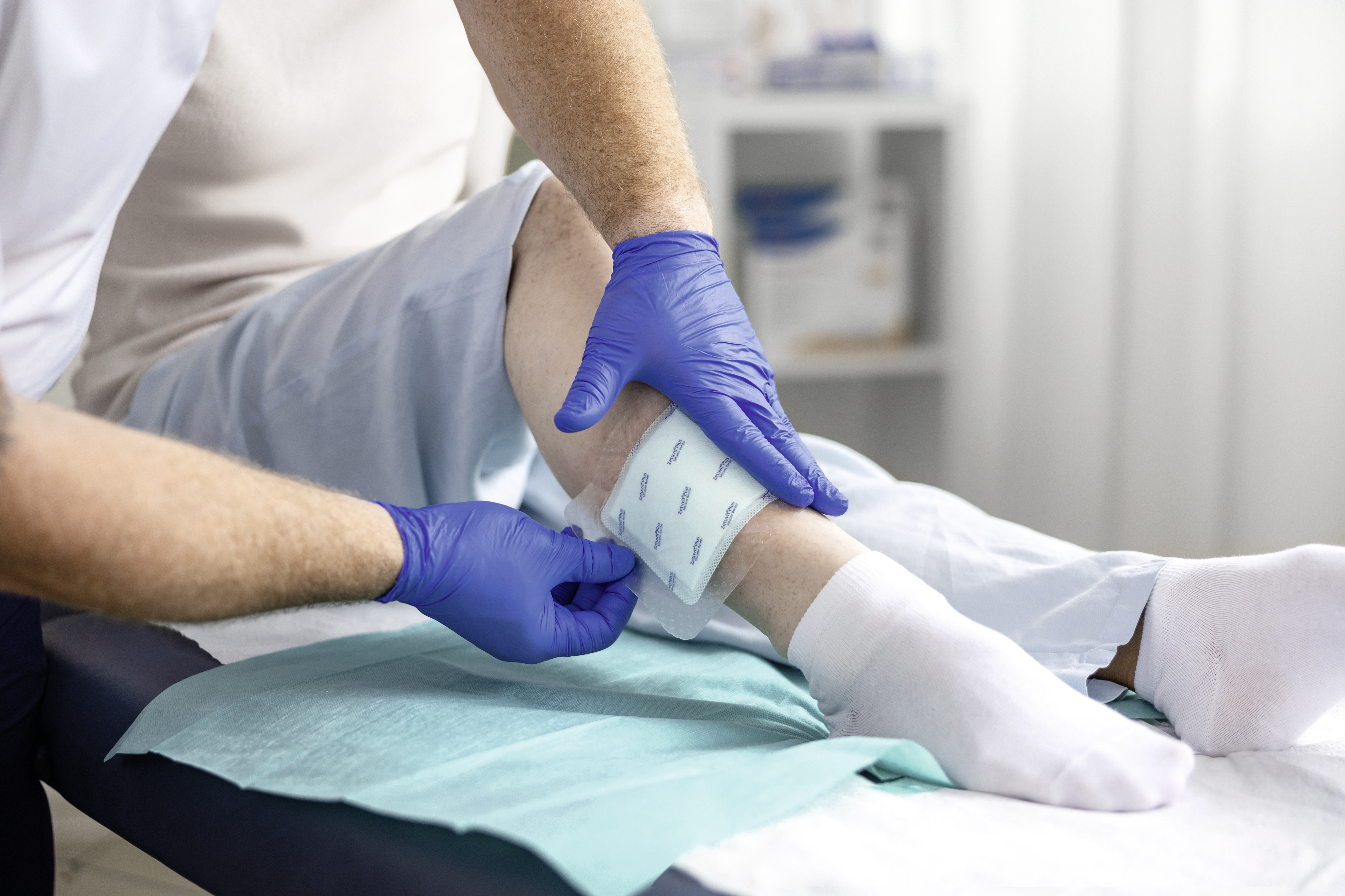Healthcare professional wearing blue gloves applies a wound dressing to a patient’s lower leg while the patient lies on an examination bed.