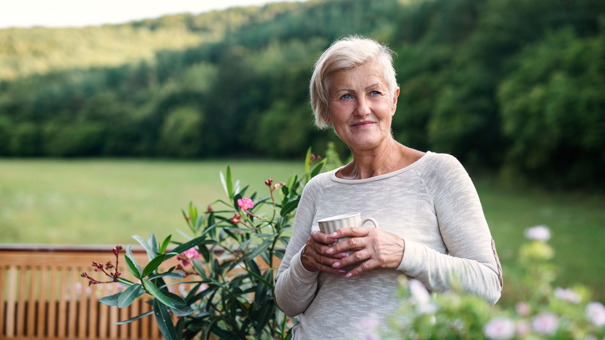Mujer de mediana edad sonriendo, tomando una infusión, sentada en un jardín.