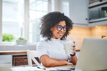 Eine Frau sitzt Zuhause an Ihrem Schreibtisch vor dem Laptop lächelnd mit einer Tasse in der Hand.