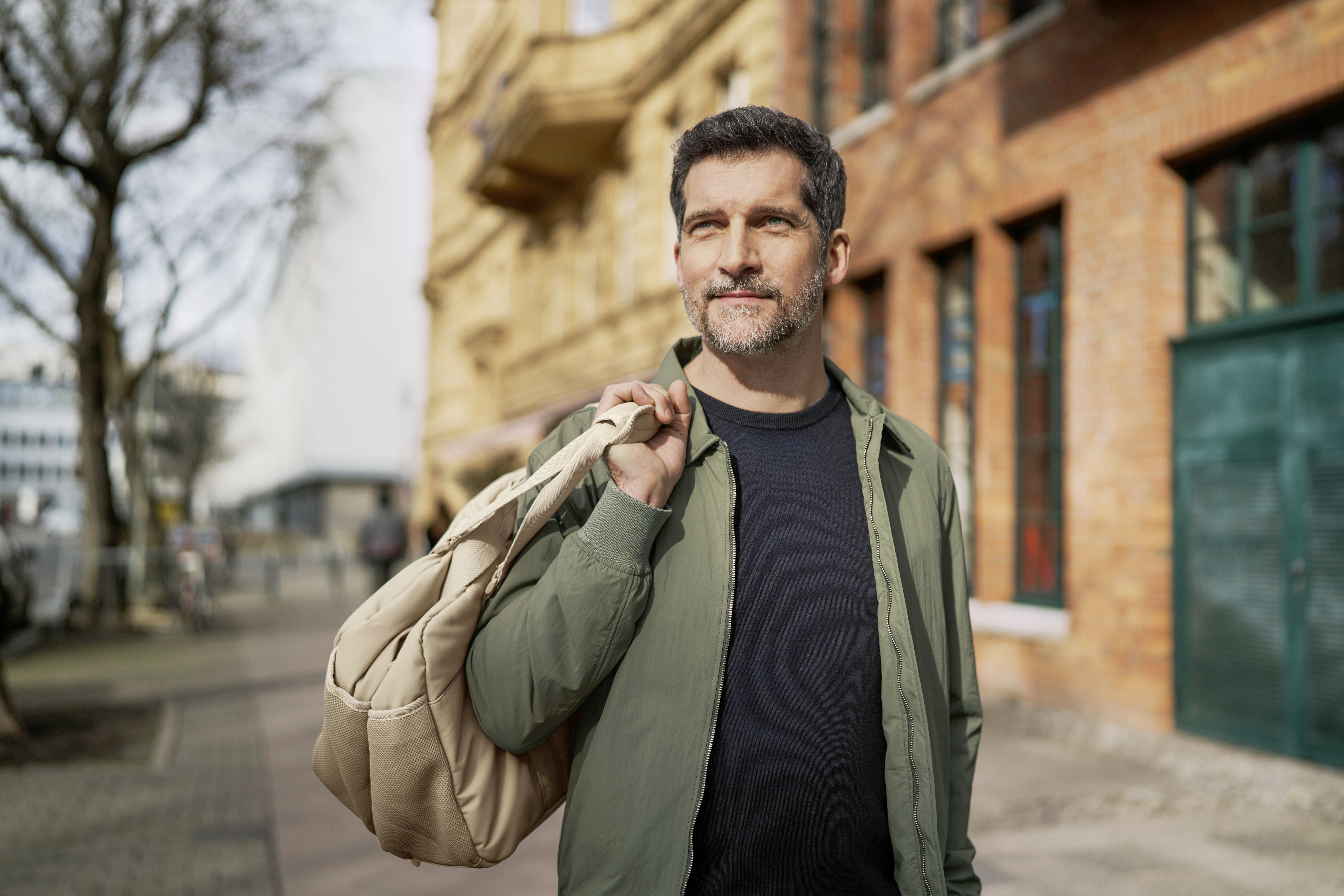Mann im schwarzen Hemd und khakifarbenen Jacke trägt eine Weekender-Tasche in urbaner Umgebung bei winterlicher Sonne.