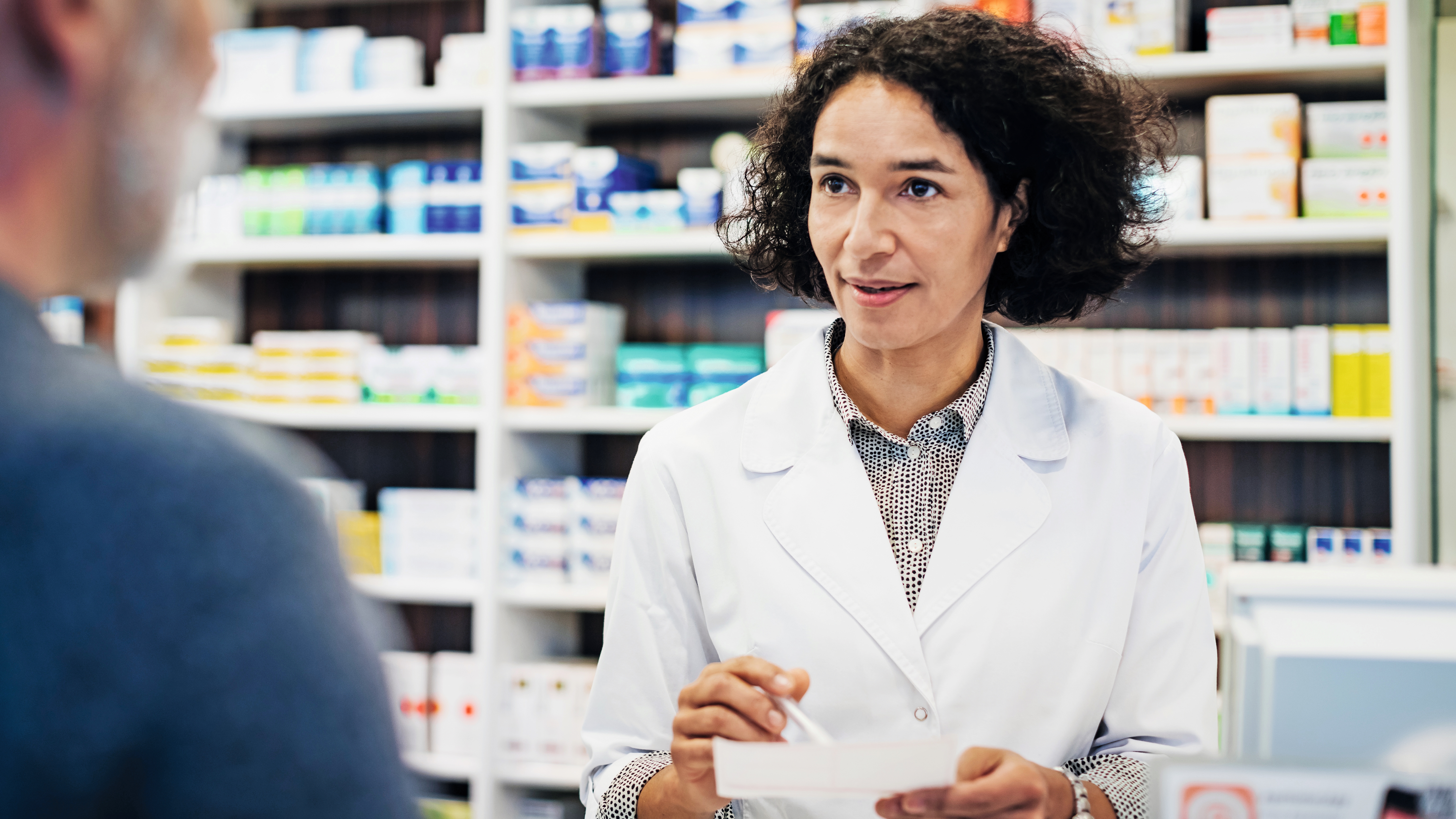 Una farmacista in camice bianco parla con un cliente mentre tiene una ricetta. Sullo sfondo, scaffali con medicinali e prodotti farmaceutici.