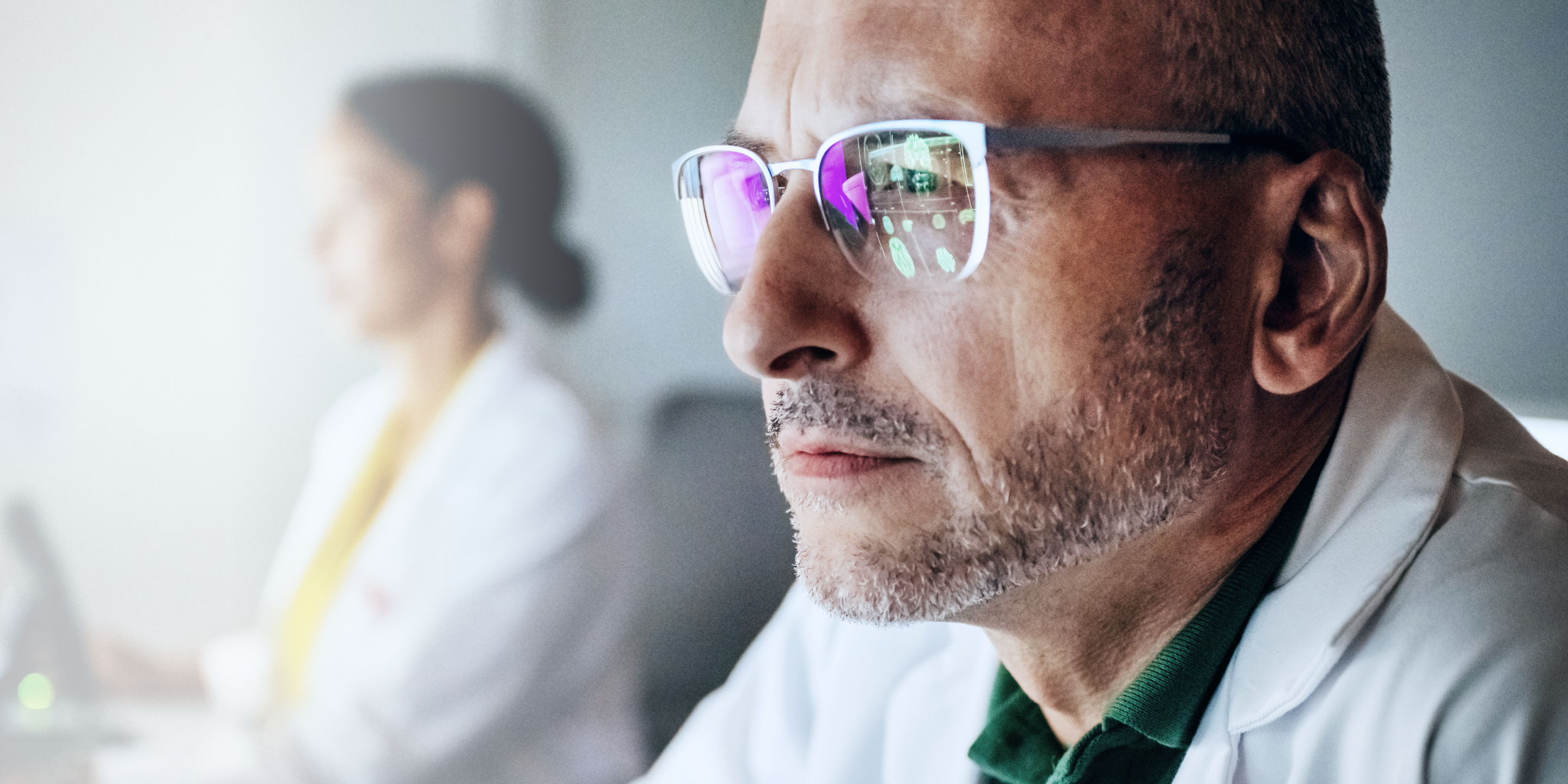 Un professionista medico concentrato, con occhiali e camice bianco, guarda con attenzione, riflettendo la dedizione in un ambiente sanitario.