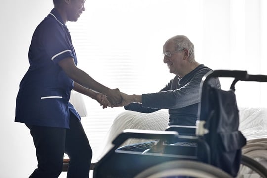 Female caregiver helping senior man get up from bed