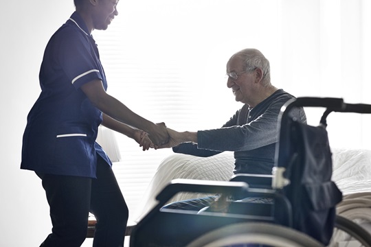 Female caregiver helping senior man get up from bed