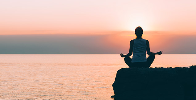 Frau in einer Yoga-Position, die im Gegenlicht in der Nähe eines Sees sitzt. 