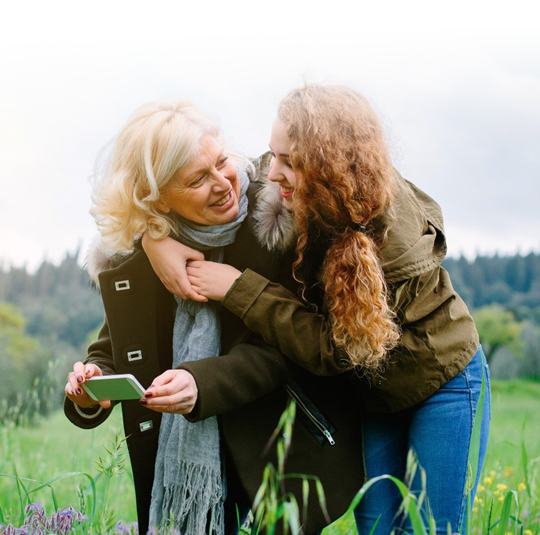 A mother and her daughter are standing on a field, smiling at each other. The daughter wrapped an arm around her mother’s shoulder, while the mother is holding a phone in her hand.