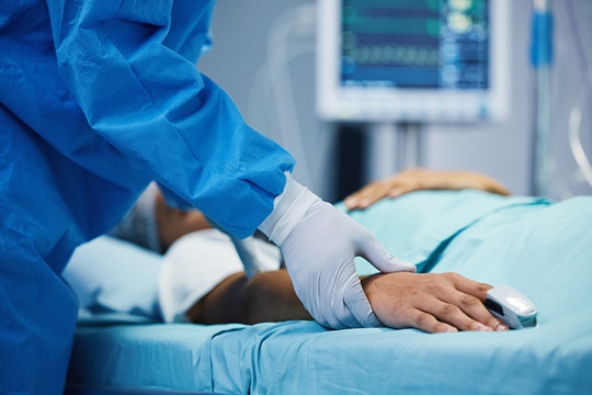 A nurse in a protective suit and gloves stands at a patient's bedside and checks their pulse.