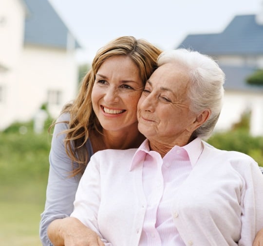 An adult woman is standing outside with her mother; both are smiling happily.