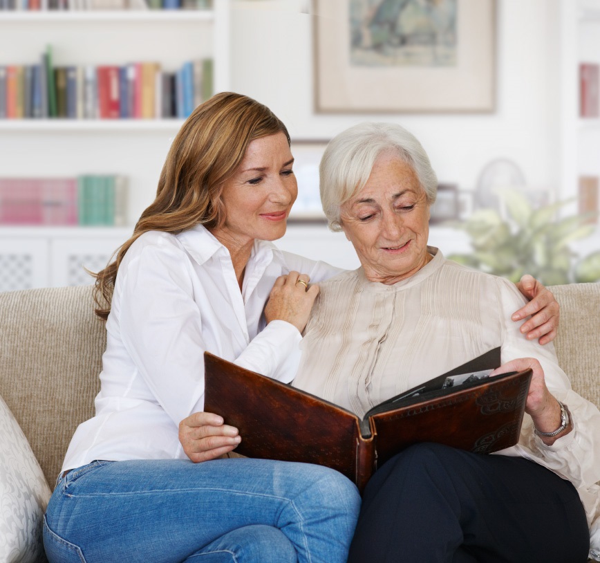 An adult woman is sitting with her mother on a sofa in a living room. The two women are looking happily at a photo album, while the daughter wrapped an arm around her mother’s shoulder.
