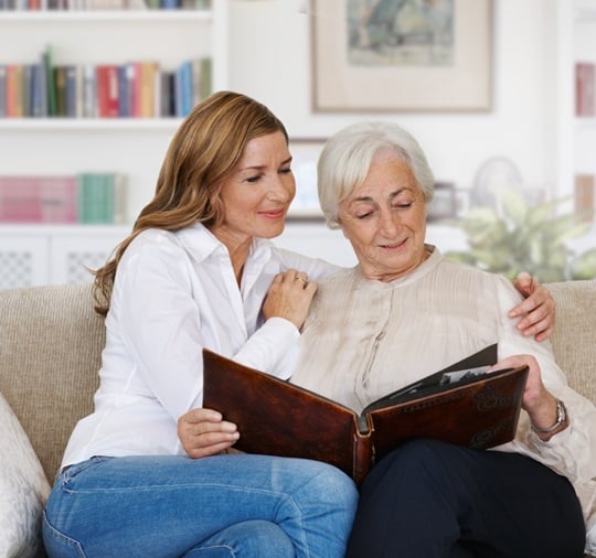 An adult woman is sitting with her mother on a sofa in a living room. The two women are looking happily at a photo album, while the daughter wrapped an arm around her mother’s shoulder.