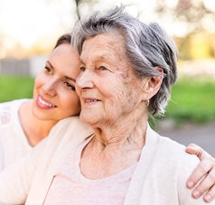 Junge Frau kniet neben Frau im Rollstuhl und hat den Arm um sie gelegt
