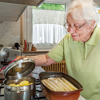 Seniorin am Herd öffnet Deckel eines Kochtopfes