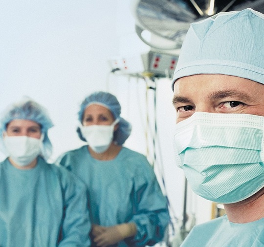 3 doctors, wearing surgical masks, are looking into the camera. Surgery equipment’s are shown in the background.