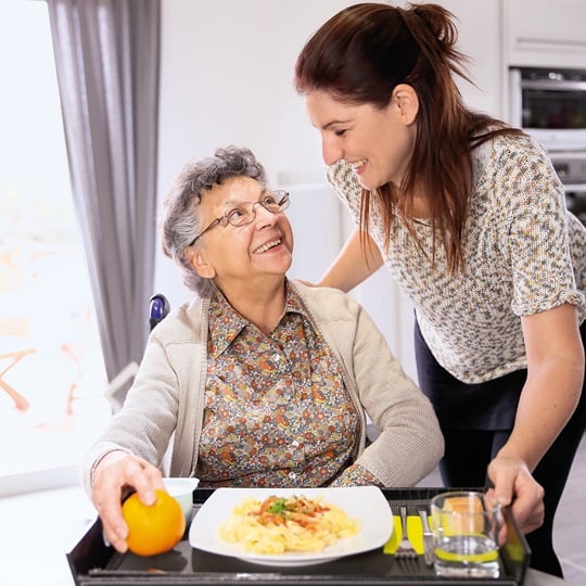 Betreuerin steht neben einer sitzenden Seniorin, die einen vollen Teller und ein Getränk vor sich stehen hat