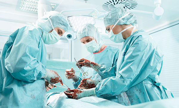 Detailný záber na operačný tím v priebehu operácie ukazuje vybavenie zdravotníckeho personálu súčasnosti, kukly, čiapky, rúšky, plášte, zástery a samozrejme chirurgické rukavice