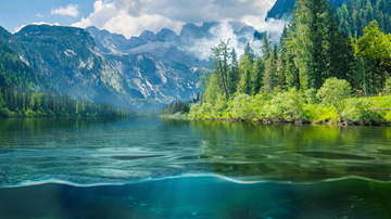 Landschaftsfoto: Im Vordergrund ein Bergsee, im Hintergrund grüne Wälder und eine Bergkette.