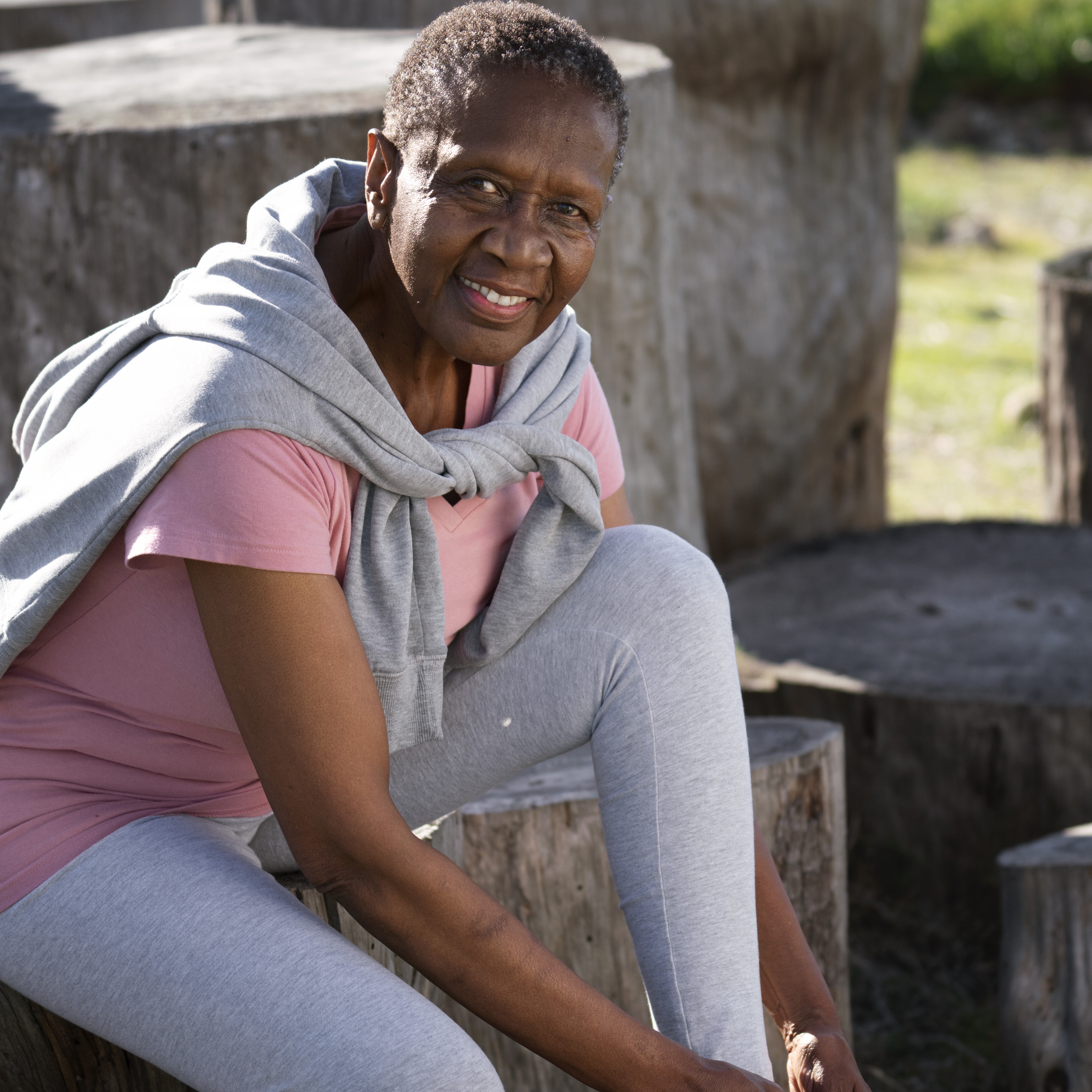 Smiling older woman in activewear tying her shoes while sitting on a tree stump outdoors.