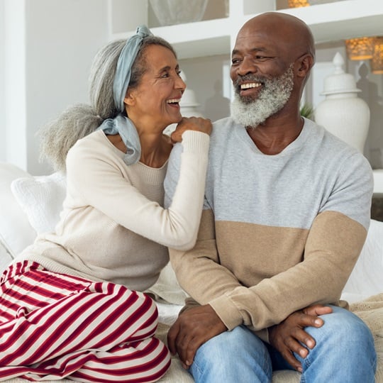 Joyful senior couple sitting together indoors, smiling at each other. The woman has her arm affectionately on the man's shoulder as they sit on a white couch in a bright, cozy room