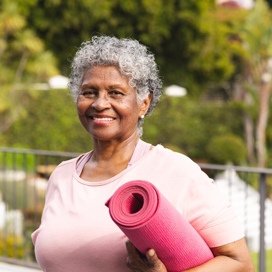Smiling senior woman with curly gray hair holding a rolled-up pink yoga mat, standing outdoors on a terrace with greenery in the background.