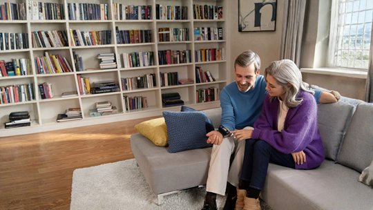 Un couple assis confortablement sur un canapé avec un dispositif de suivi de la santé, entouré d’une bibliothèque chaleureuse.