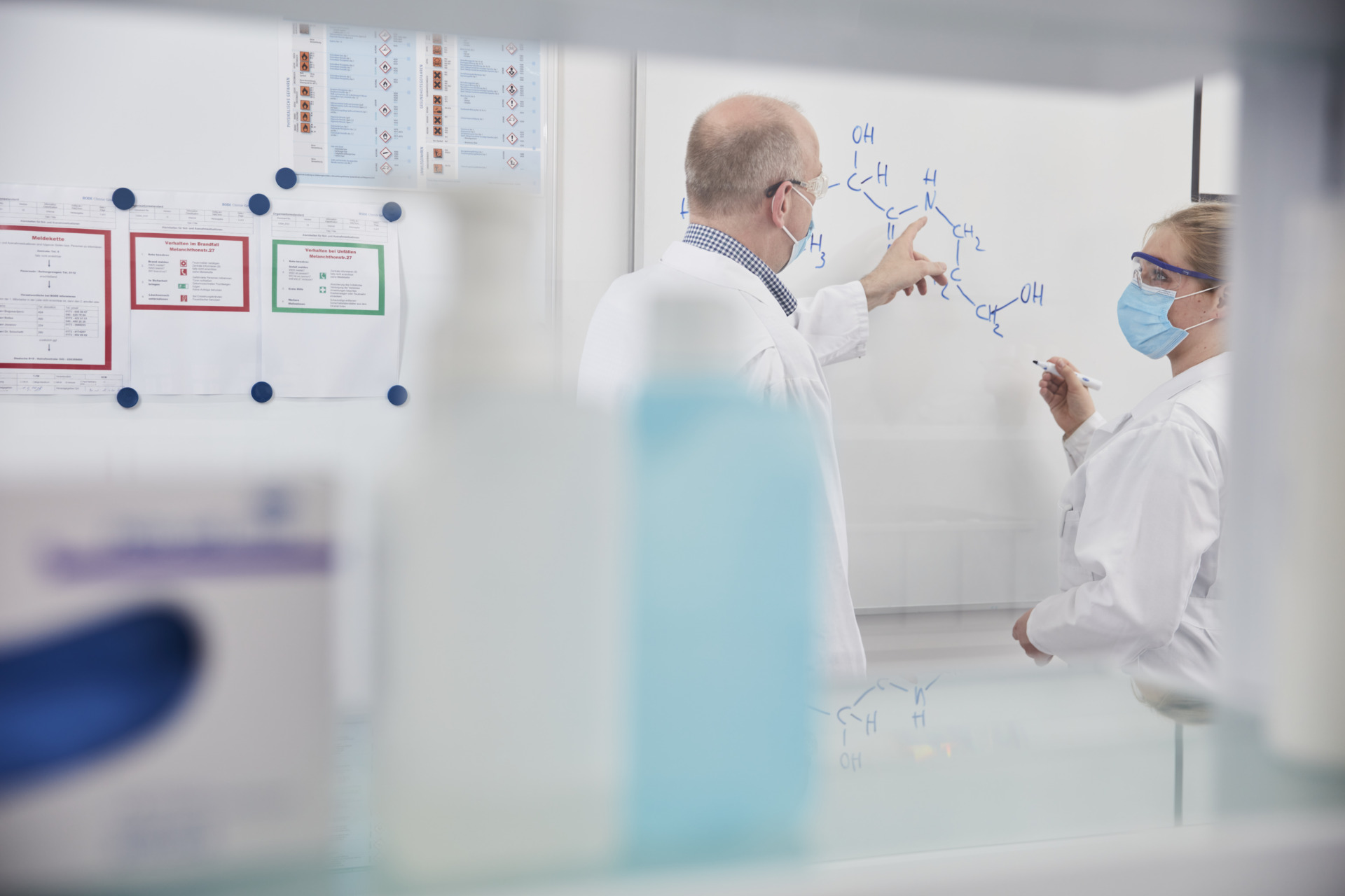 Zwei Wissenschaftler in Laborkitteln und Masken analysieren Informationen auf einem Whiteboard in einem Forschungslabor.