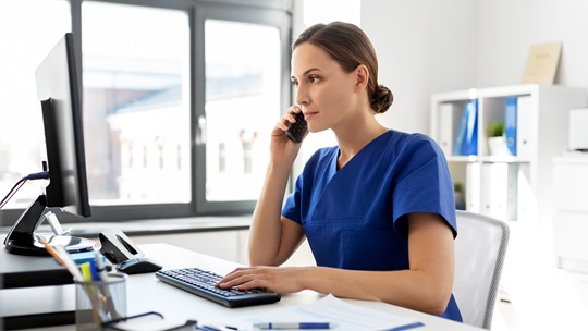 Pflegekraft sitzt vor dem PC und Telefoniert mit einem Mobiltelefon