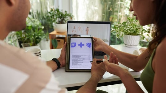 Mann und Frauen sehen gemeinsam in ein Tablet. Im Hintergrund ist ein Laptop zu sehen