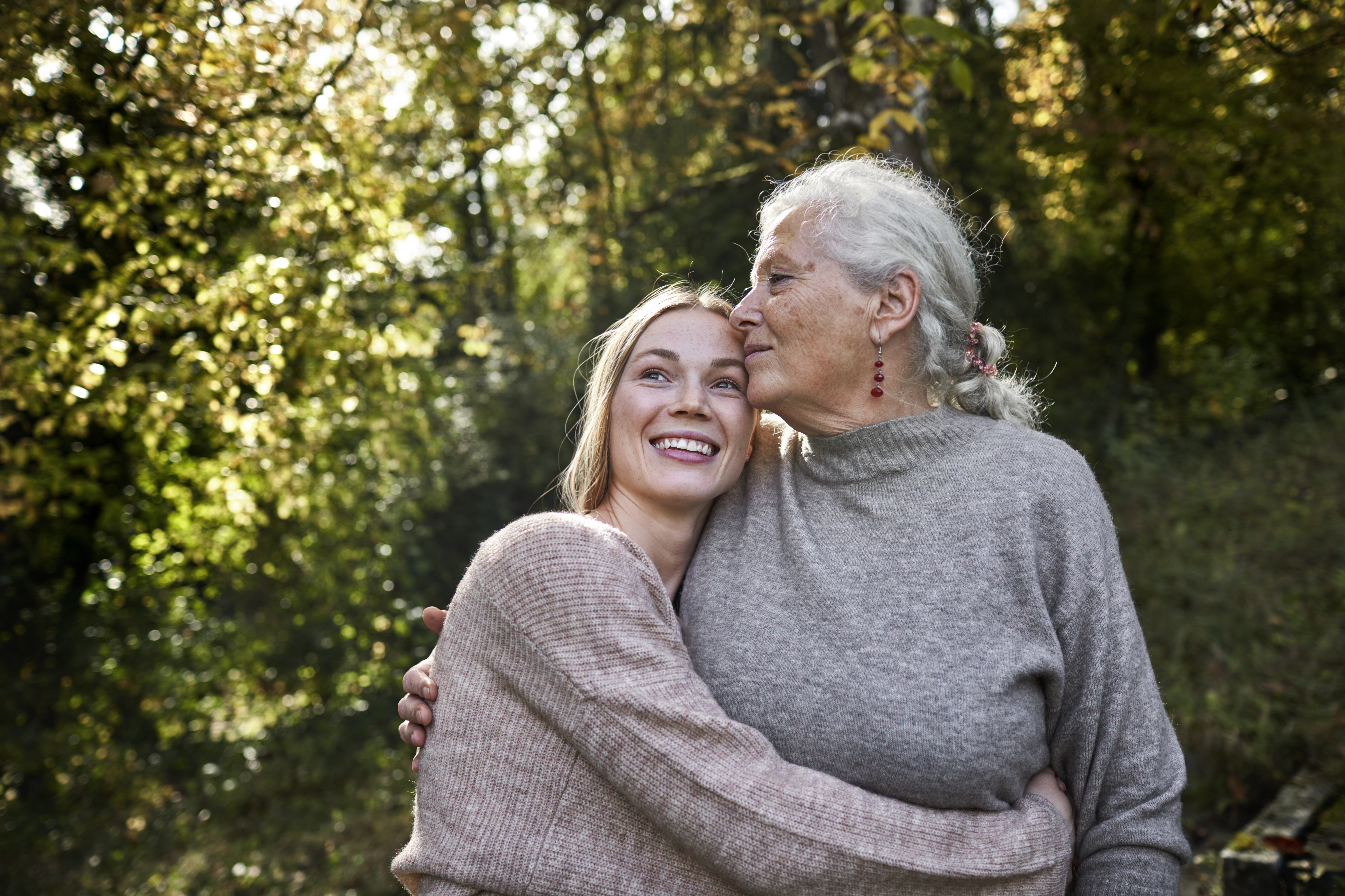 An elderly woman with gray hair and red earrings lovingly kisses the forehead of a smiling younger woman, while they embrace outdoors in a sunlit forest setting. Both are wearing cozy sweaters, creating a warm and affectionate scene.
