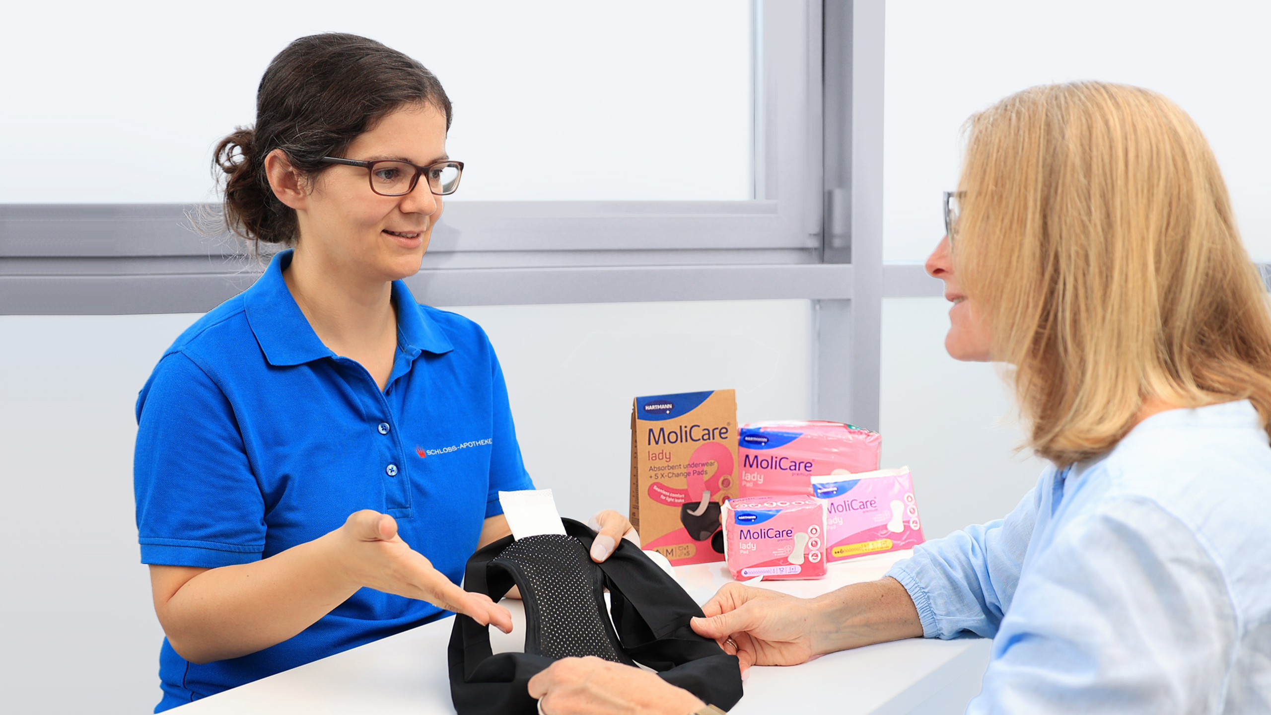 Apothekerin in blauem Poloshirt präsentiert einer Kundin verschiedene MoliCare Produkte.