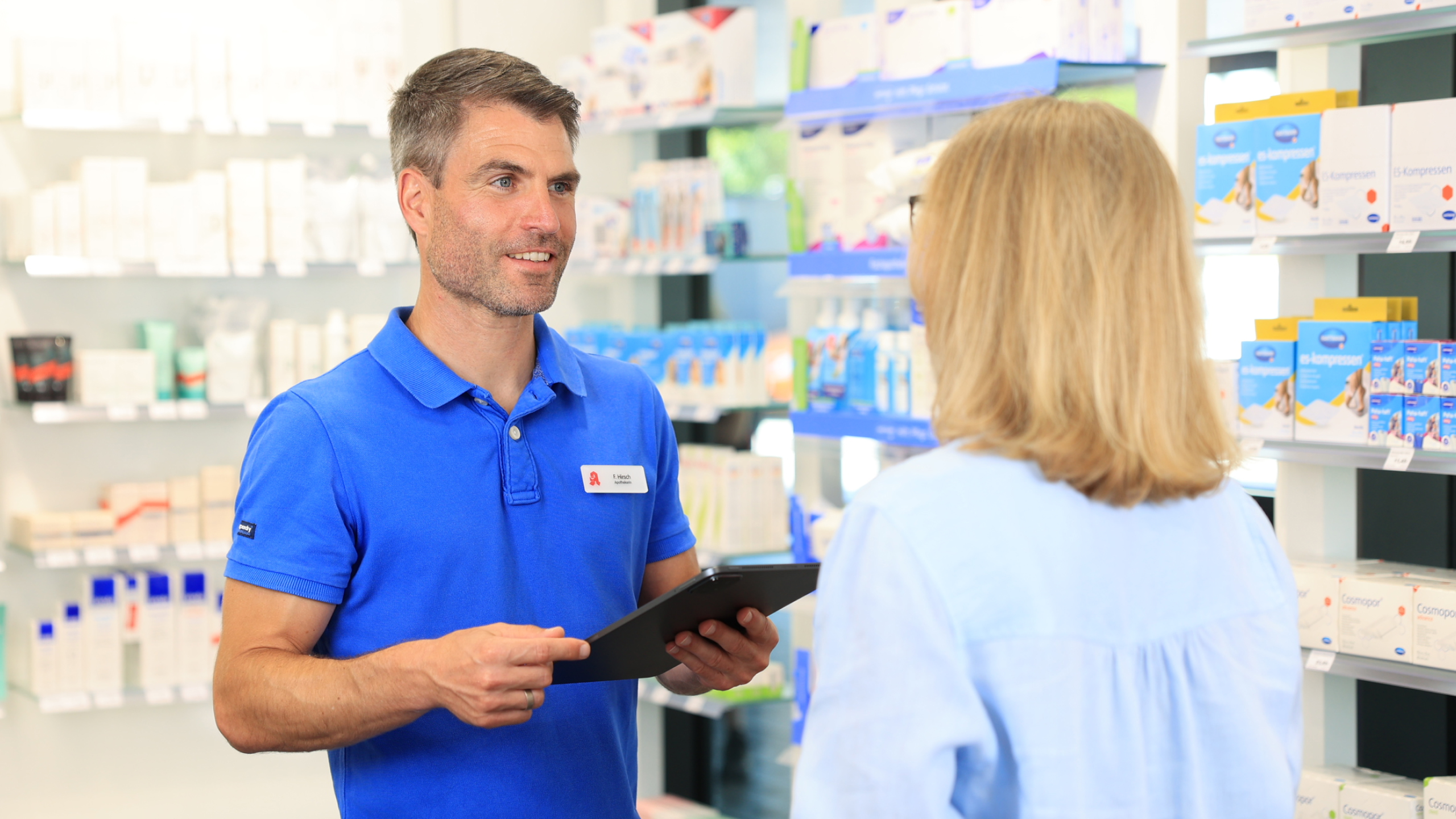 Apotheker in blauem Poloshirt und Tablet in der Hand im Gespräch mit einer Kundin.