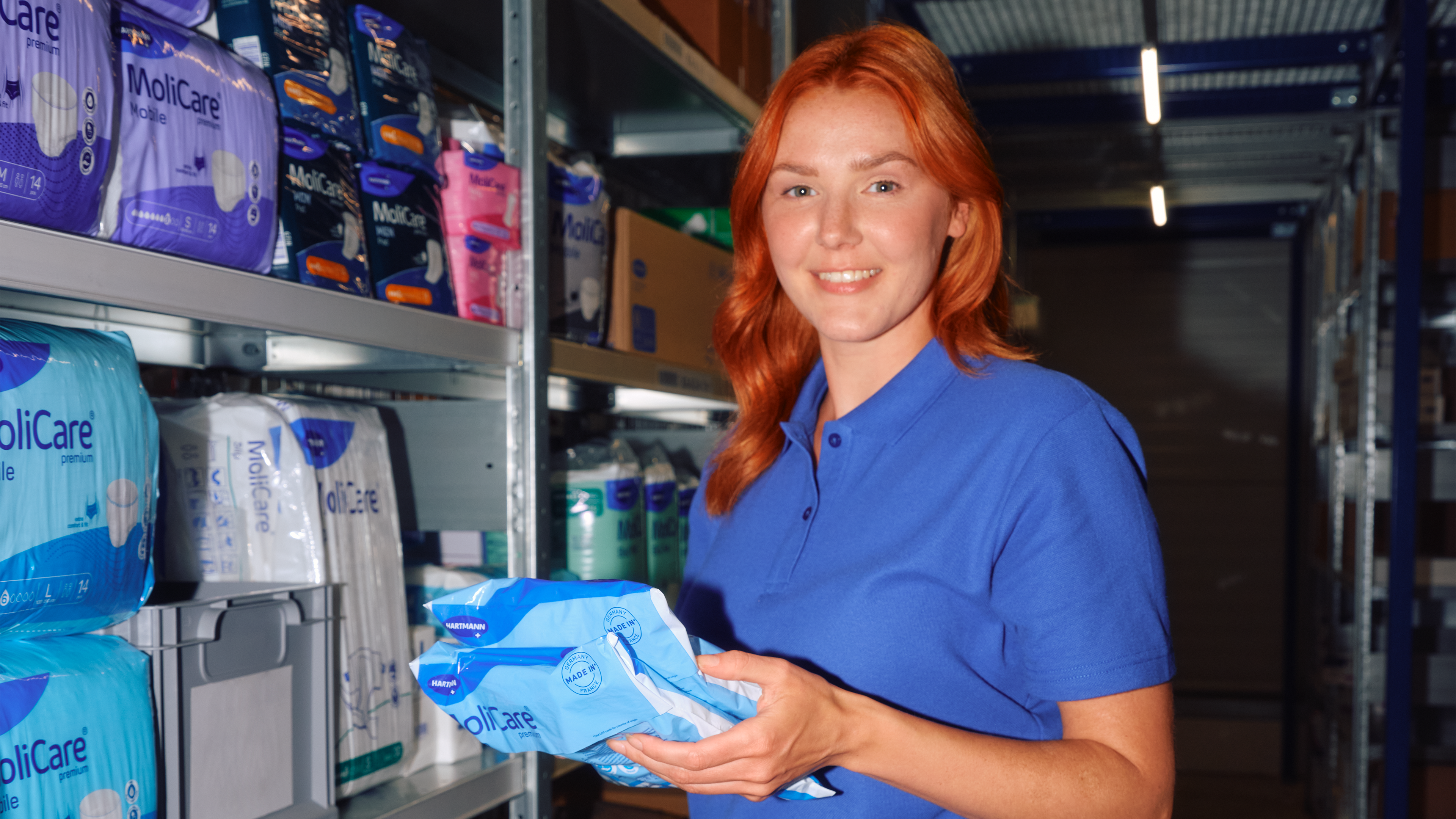 Person in blauem Poloshirt hält MoliCare Produkte vor einem Lagerregal mit Hygiene- und Inkontinenzartikeln. PHI26_01_14