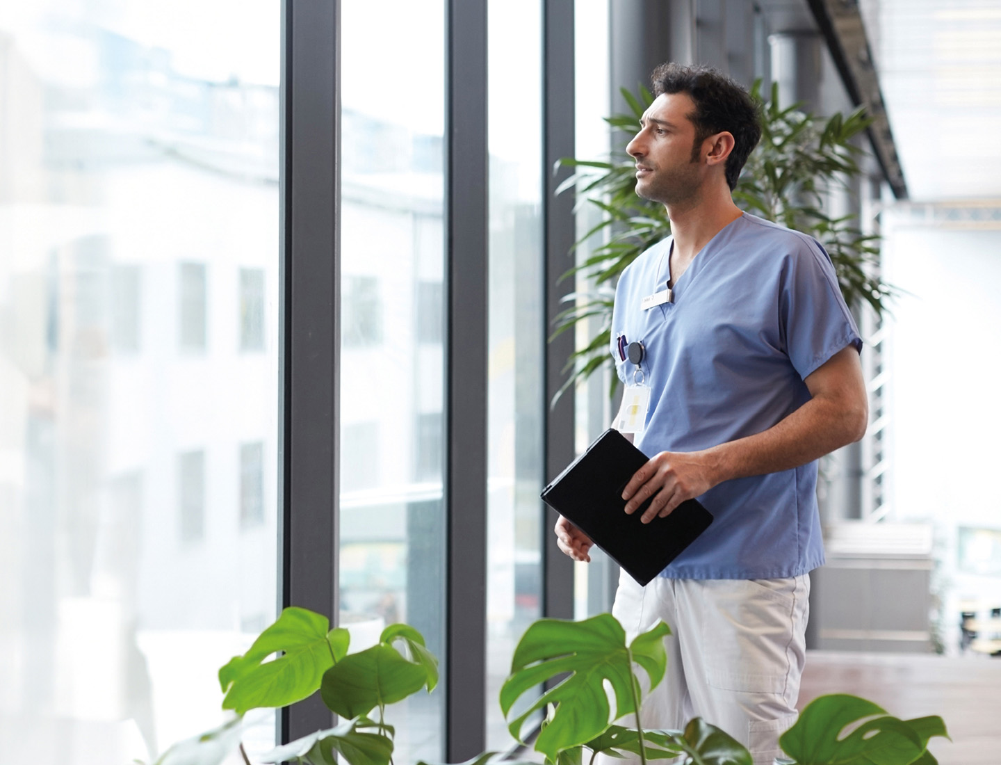 Eine medizinische Fachkraft in blauem Shirt und weißer Hose steht entspannt an einem Fenster mit Pflanzen. In der Hand hält sie ein Tablet – ruhig, professionell und zuversichtlich.