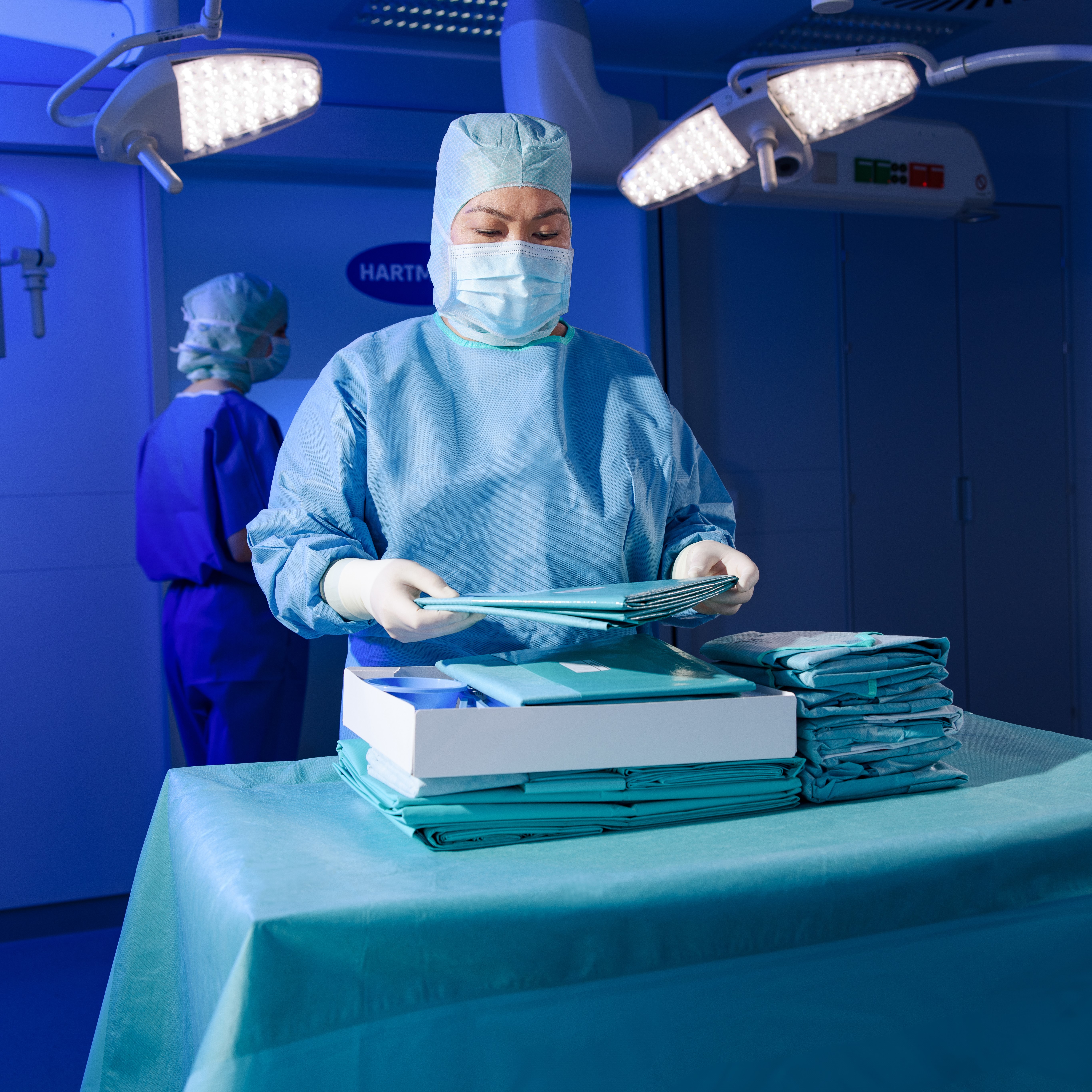 OP-Fachkraft im sterilen Mantel und Mundschutz sortiert Abdecktücher in einem hell ausgeleuchteten OP-Raum – mit Fokus auf Präzision und Hygiene.