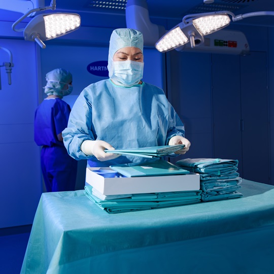 Medical professional in a surgical gown and mask organizes sterilized drapes in a well-lit operating room setting, focusing on precision and hygiene.