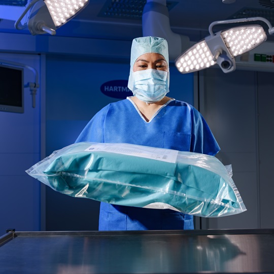 Medical professional in surgical attire holds a sealed sterile package under bright operating room lights.