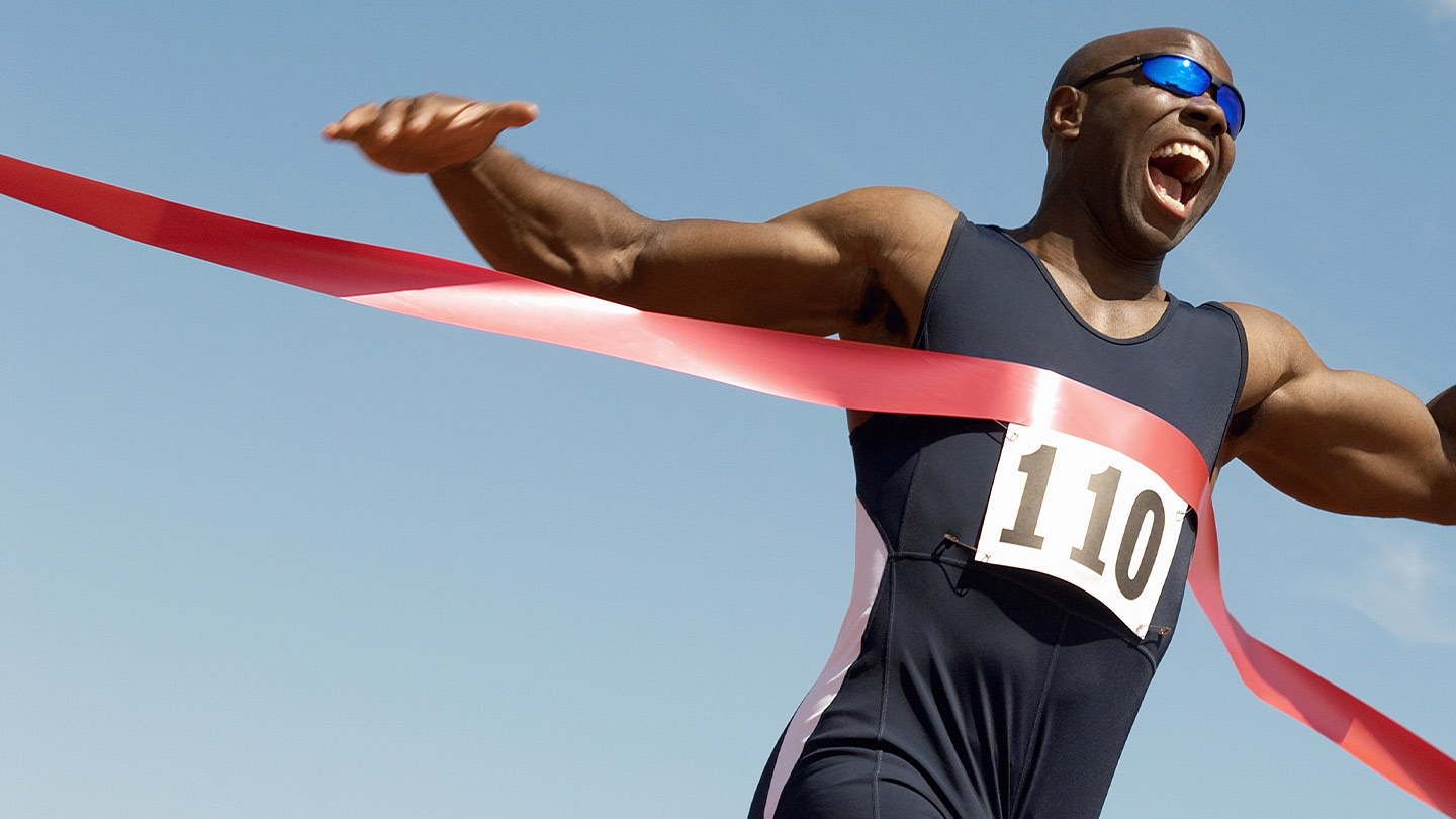Male sprinter triumphantly crossing the finish line with arms raised and mouth open in celebration, wearing race number 110 and sunglasses under a clear blue sky.