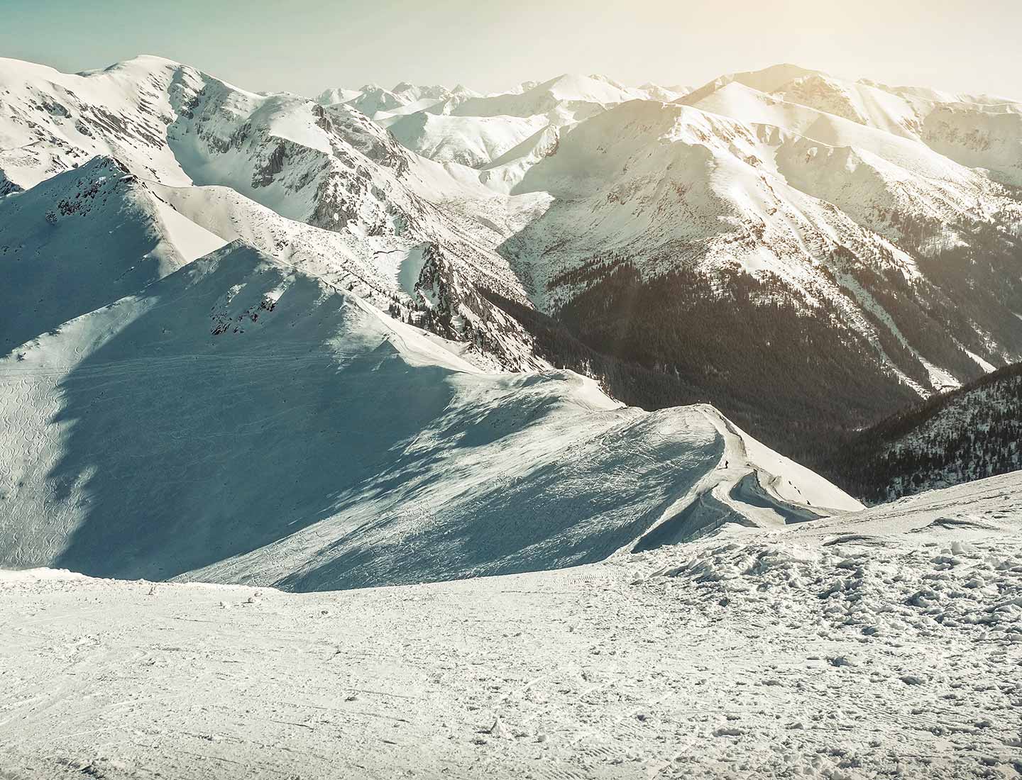 Panoramic view of snow-covered mountain range and ski slopes under a clear sky, with traces of skiing visible in the snow.