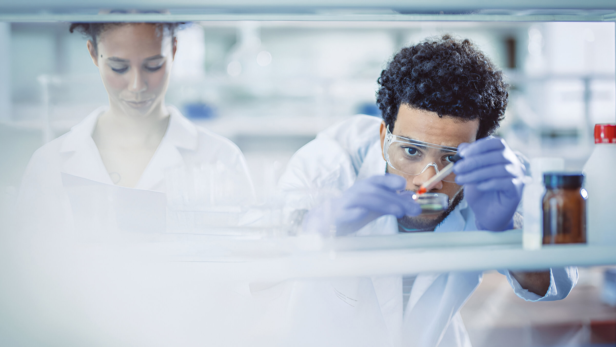 A scientist wearing protective goggles and gloves examines a Petri dish.