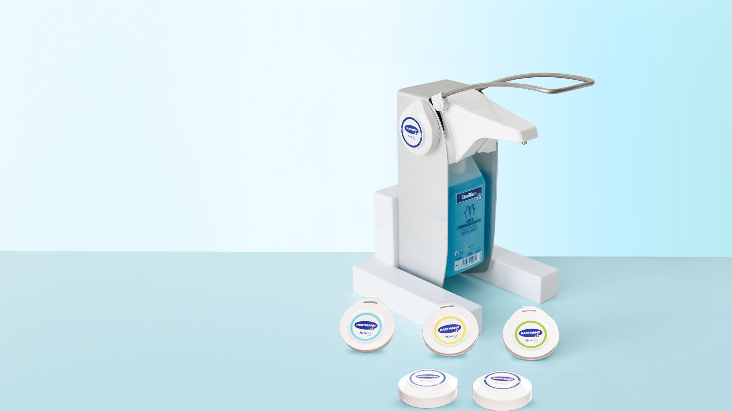 A white Sterillium® hand sanitizer dispenser with a blue liquid container, positioned on a light blue surface, surrounded by four white refill pods labeled with different colors.