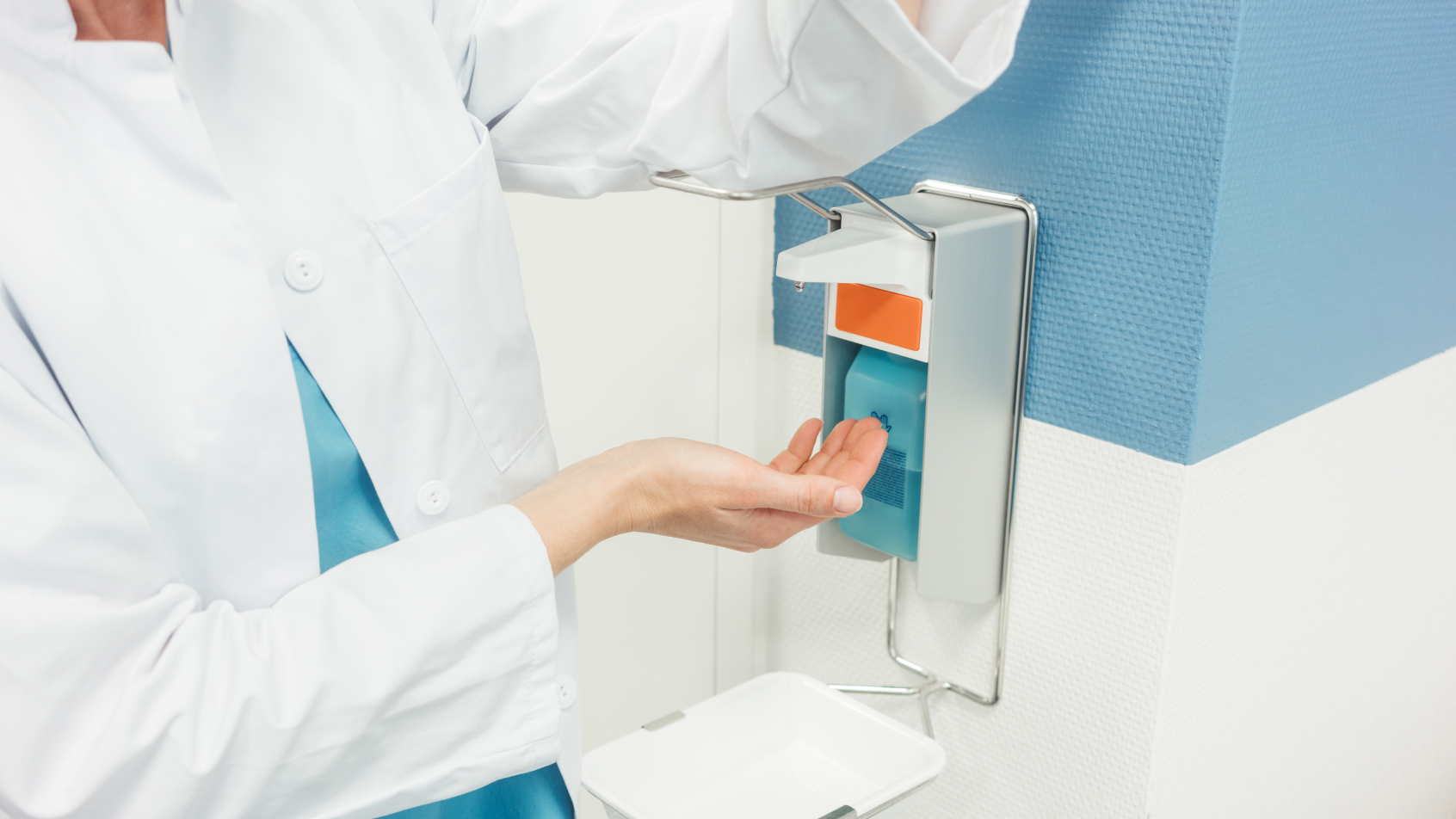 A doctor uses a hand sanitizer dispenser by pressing on the lever with the left elbow and catching the disinfectant in the palm of their right hand.