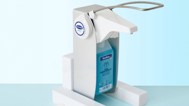 A white Sterillium® hand sanitizer dispenser with a blue liquid container, positioned on a light blue surface, surrounded by four white refill pods labeled with different colors.