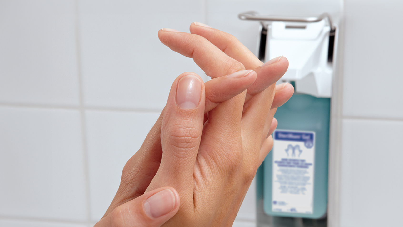 Set of hands applying Sterillium® in front of a wall mounted metal dispenser for Sterillium®.