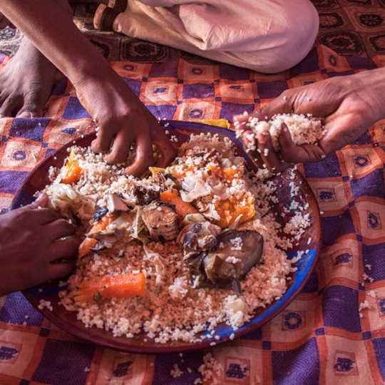 Three people eat with their hands from a bowl of couscous with vegetables on the floor between them.