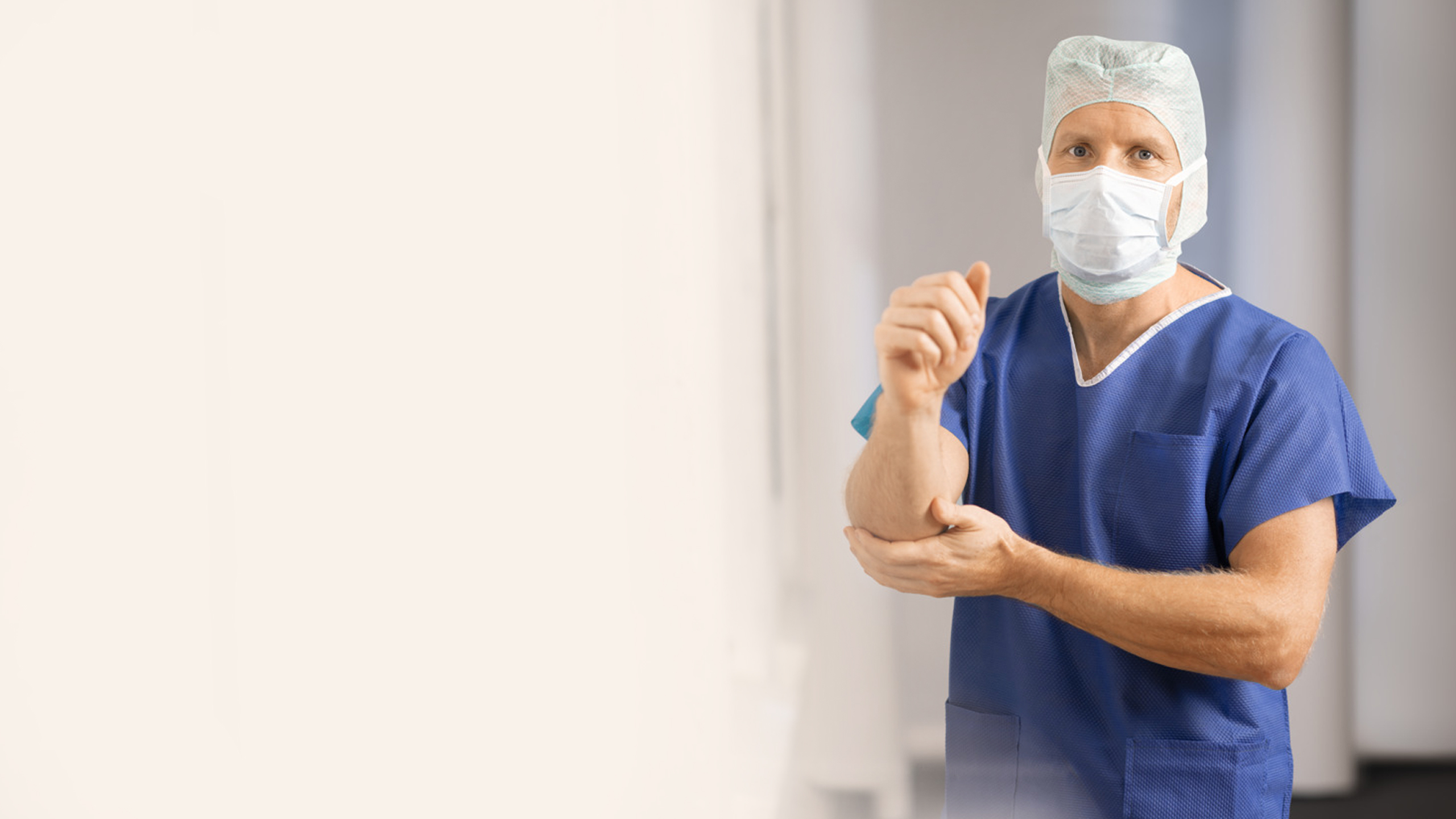 A doctor wearing a facemask, a medical hood, and blue scrubs disinfects his hands and arms to enter the operation room.