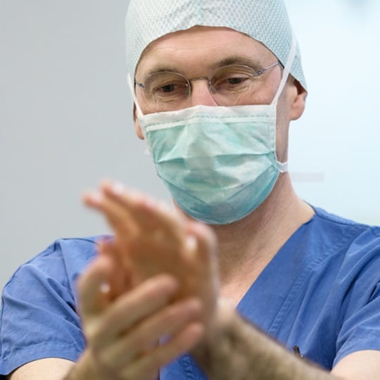 A male doctor wearing a facemask, a pale blue surgical hood, and blue scrubs disinfects his hands.