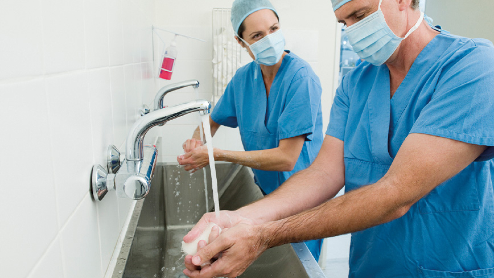 Two doctors in facemasks and scrubs wash their hands before surgery.