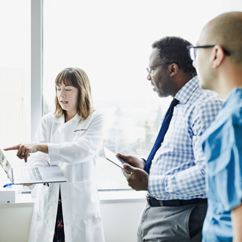 A female doctor in a white coat shows something on a laptop screen to a man in a suit and a young nurse.