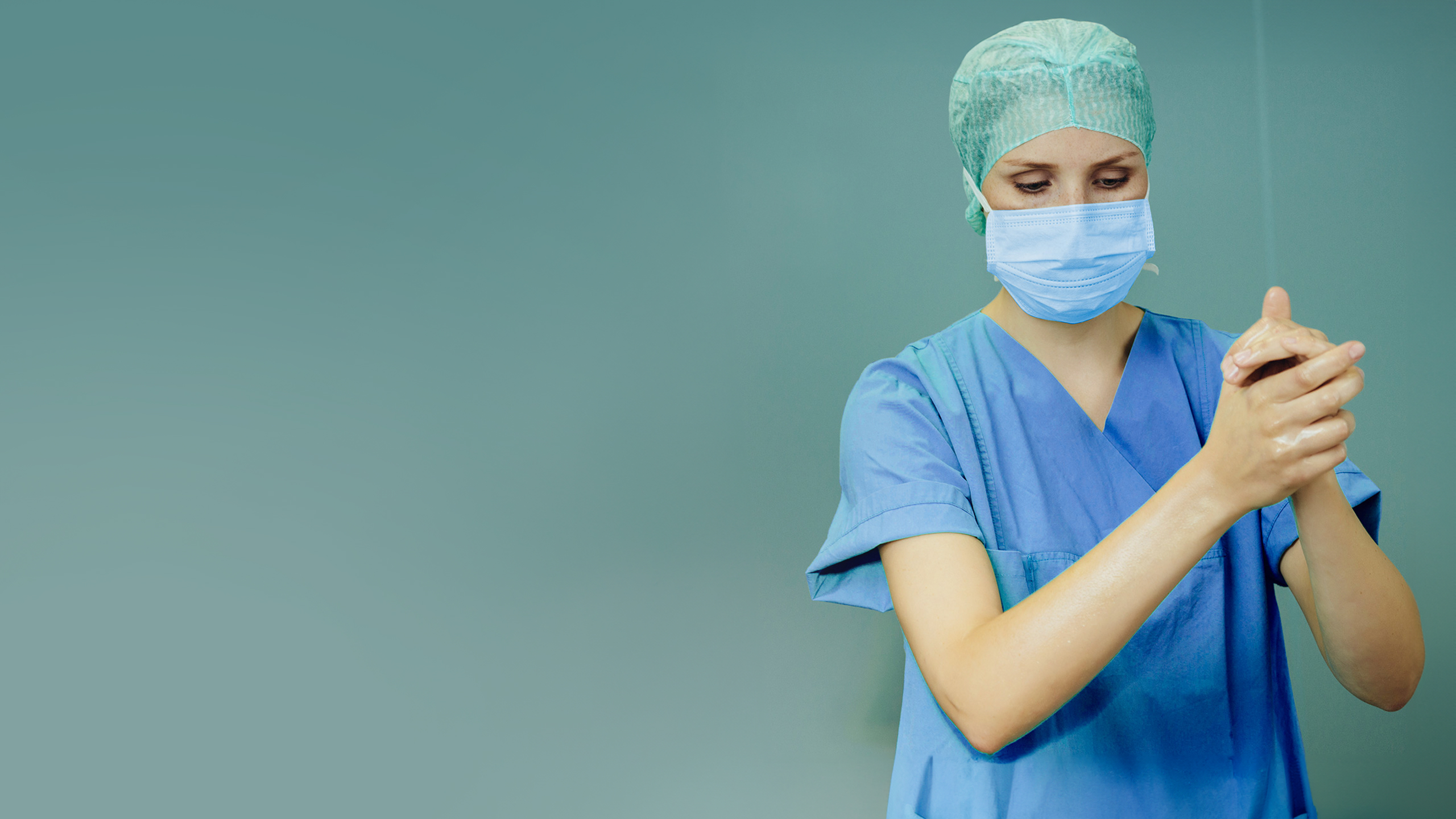 A female doctor wearing a facemask, blue scrubs, and a green surgical hood disinfects her hands.