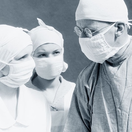 Schwarz-Weiß-Foto eines Arztes und zweier Krankenschwestern, die Kleidung aus dem frühen 20. Jahrhundert und Gesichtsmasken tragen.