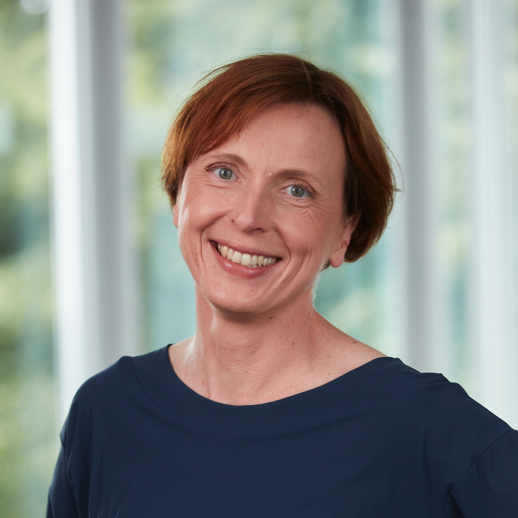 Portrait of Dr. Heide Niesalla, Head of HARTMANN SCIENCE CENTER (HSC), in front of a bright, blurred background with large windows. She is wearing a dark blue top and smiling at the camera.